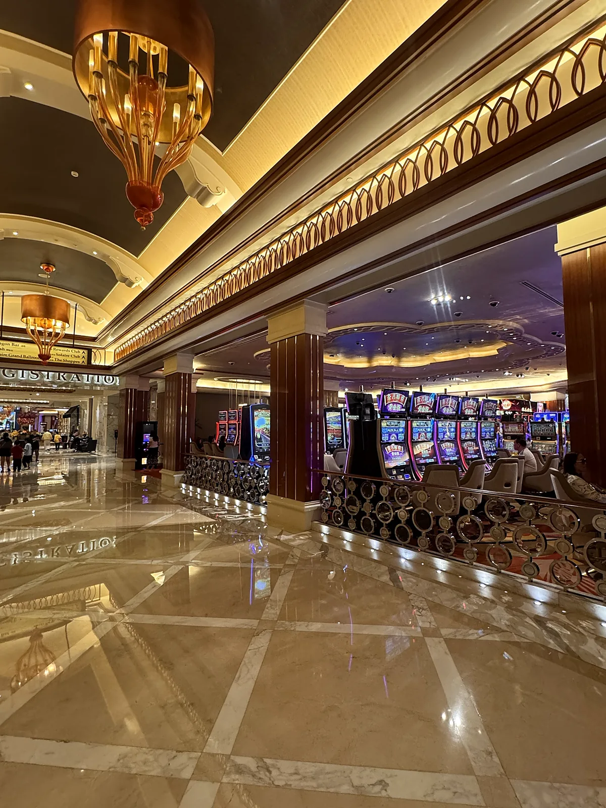 Casino gaming floor with rows of slot machines beside a marble lobby corridor lit by amber glass chandeliers