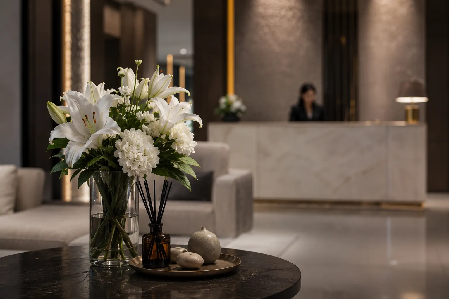 Reed diffuser and a white lily arrangement on a lobby table, with a hotel reception desk softly out of focus behind
